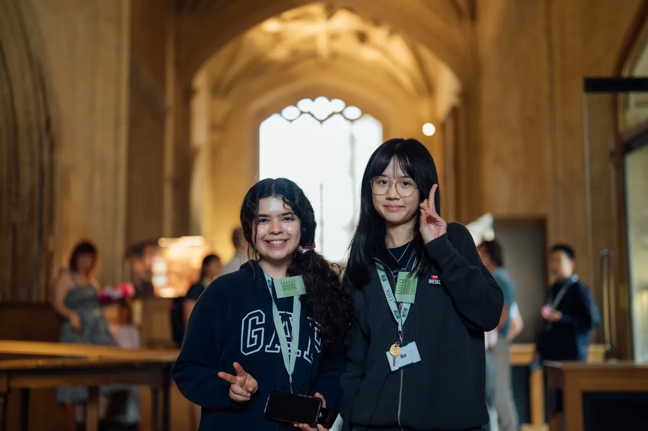 Two smiling young women indoors, one holding a phone, both wearing lanyards with name tags.