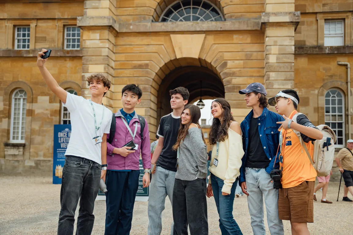 Group of six young people posing for a selfie outside a historic stone building with arched entrance.