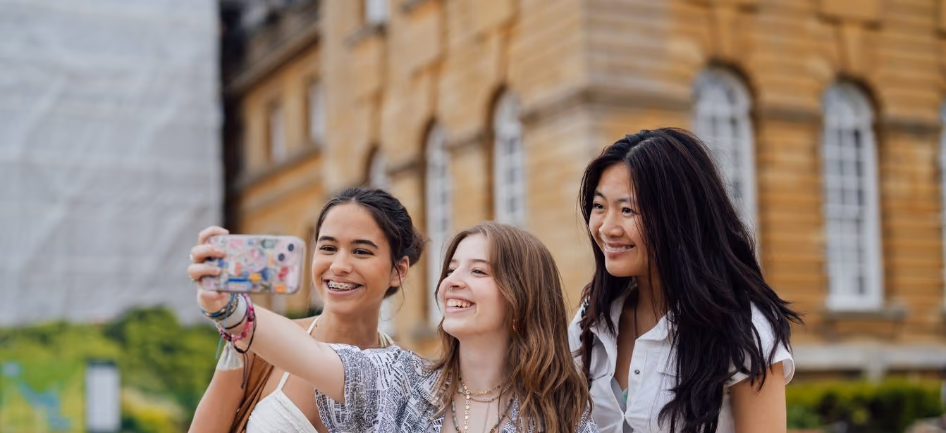 Three young women smiling and taking a selfie outdoors with historic building in background.