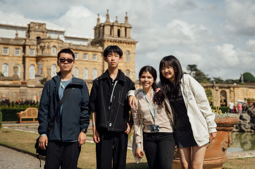 Four young people posing smiling outdoors in front of a historic stone building under a cloudy sky.