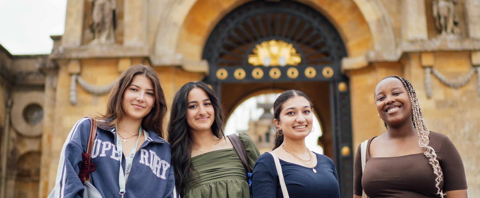 Four diverse young women smiling and standing together outdoors in front of an ornate archway.