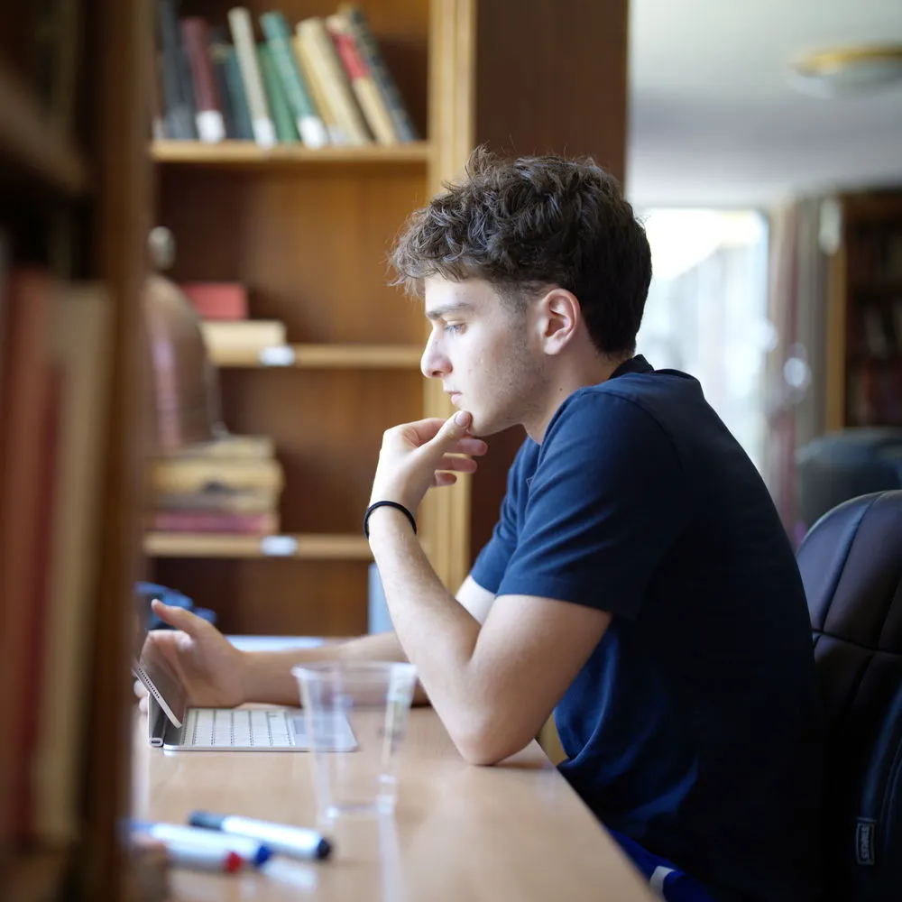 Young man sitting at desk inside library reading a tablet with a thoughtful expression.