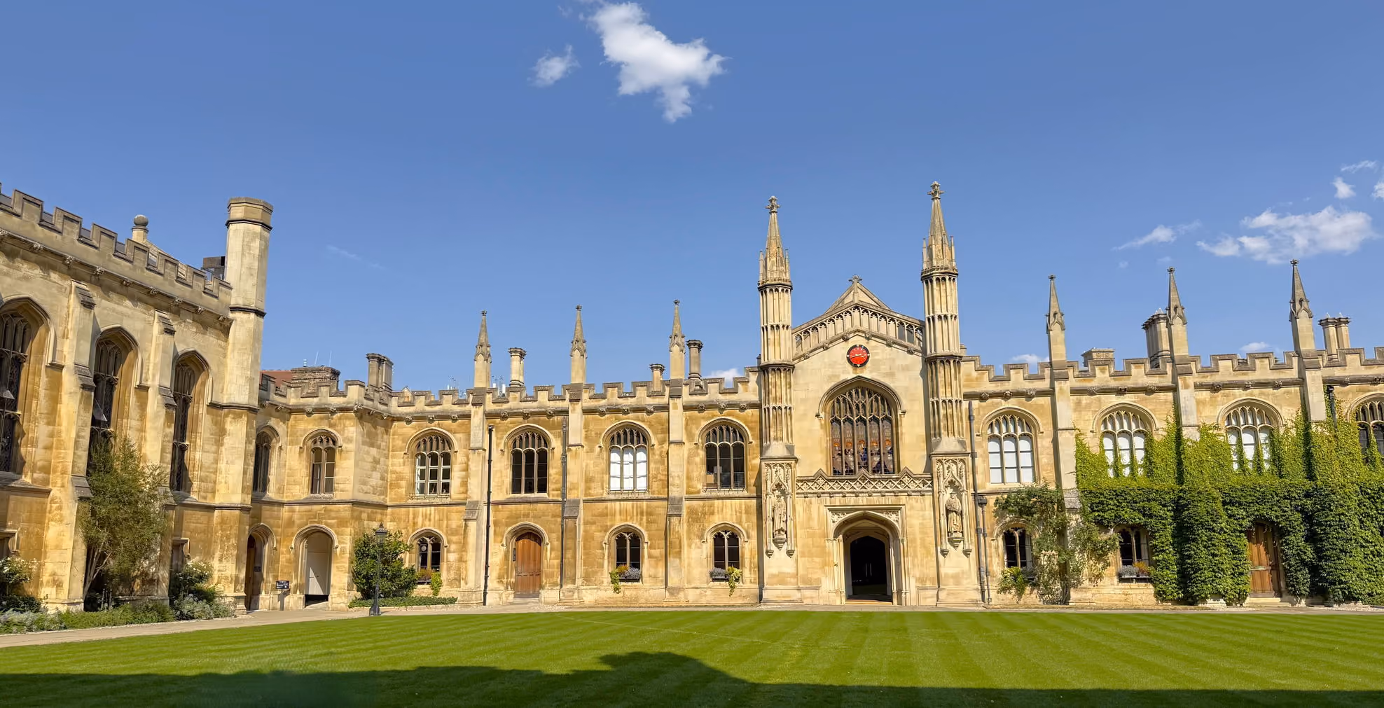 Historic stone building with arched windows, spires, and a green lawn under blue sky.