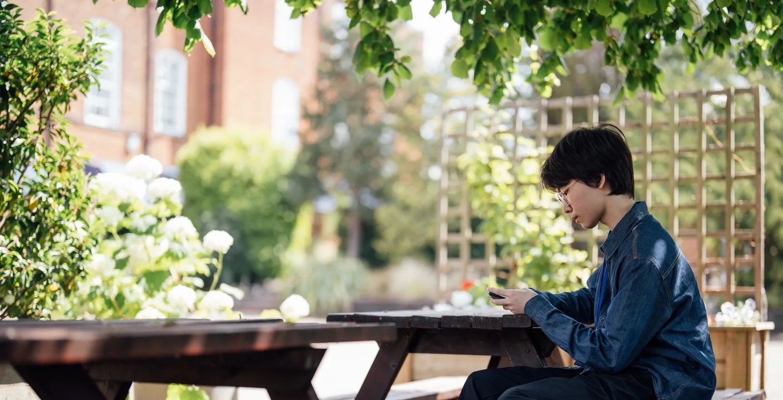 Person in glasses and denim jacket sitting at picnic table outdoors, looking at their phone.