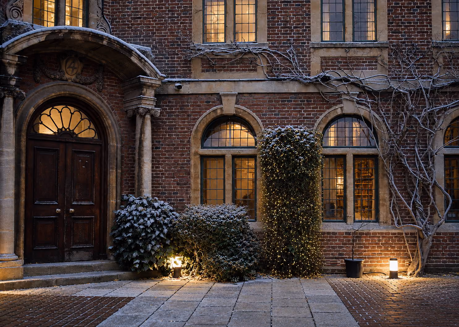 Brick building facade with arched windows, wooden door, lit bushes, and a cobblestone courtyard at dusk.