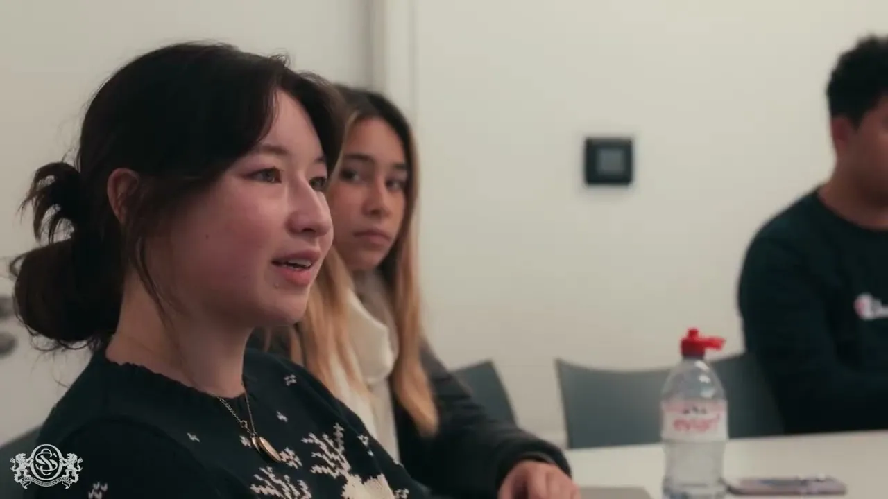 Three people sitting at a table in a meeting room, one woman speaking and others listening.