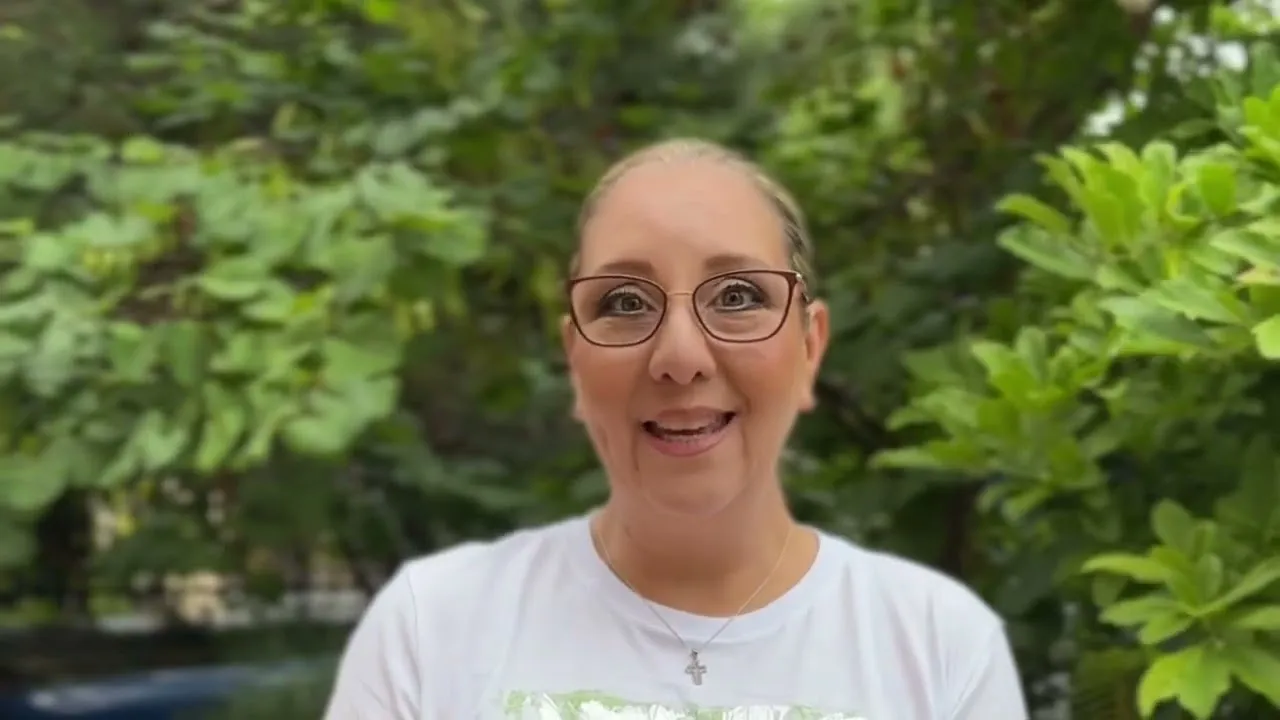 Smiling woman with glasses wearing a white shirt and cross necklace standing in front of green foliage.