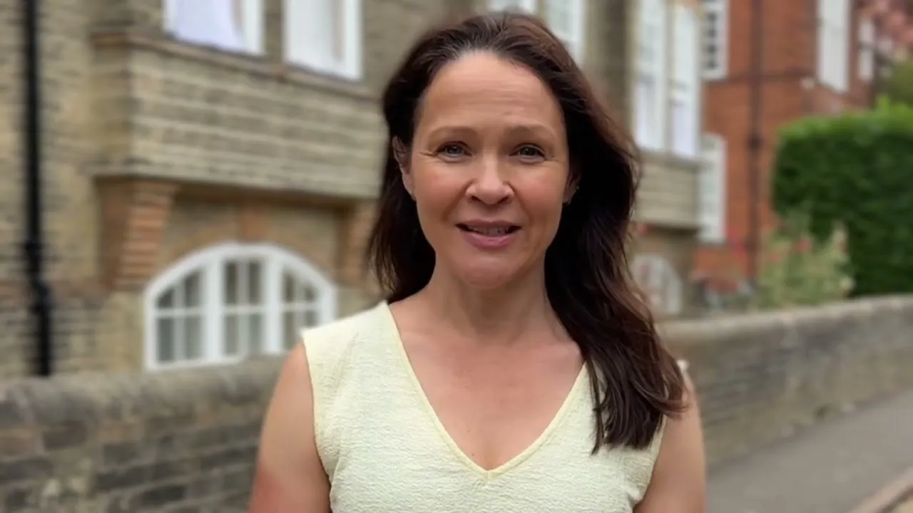 A woman with long brown hair smiles while standing outdoors near a brick wall and buildings.