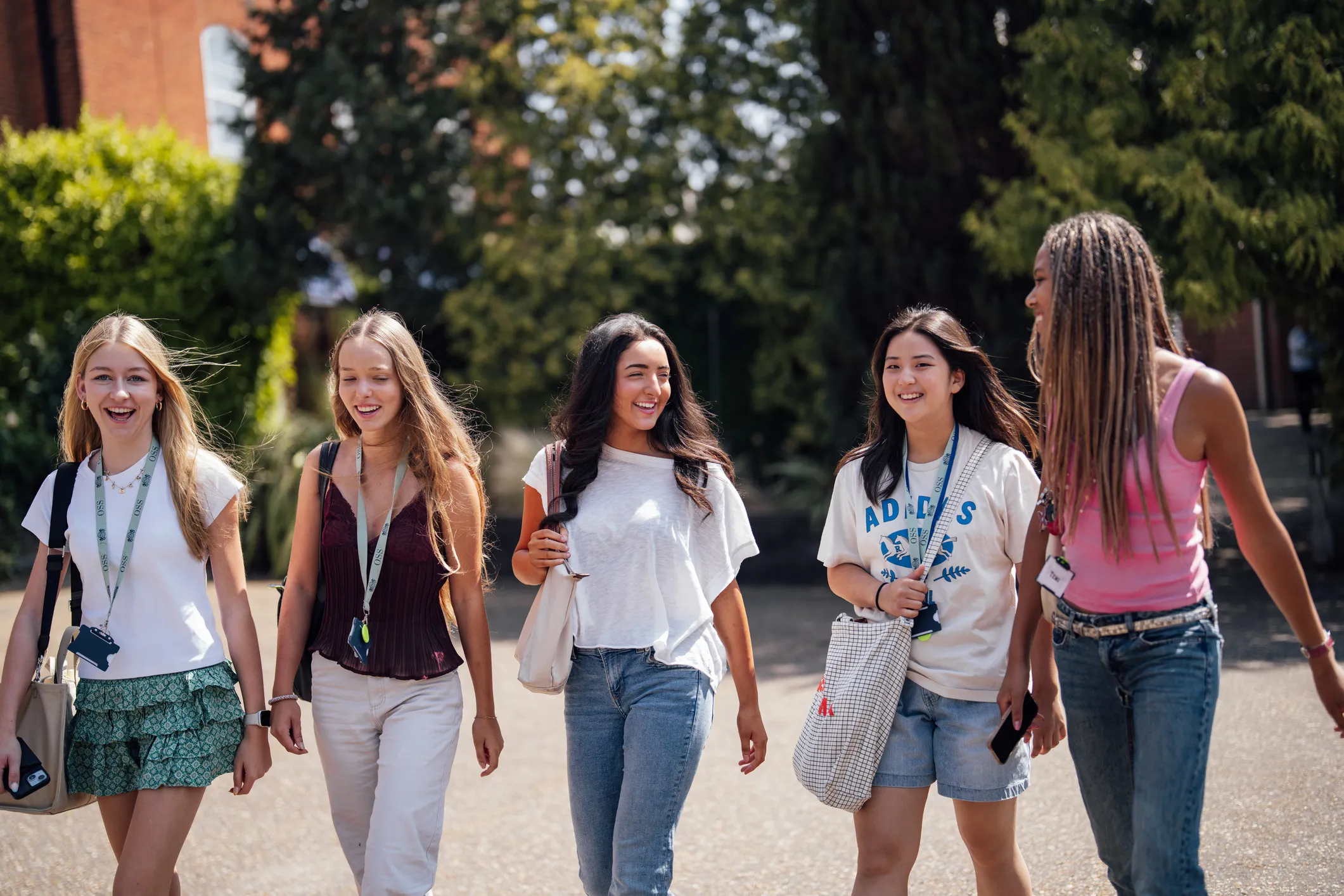Five young women walking outdoors on a sunny day, smiling and carrying bags and phones.