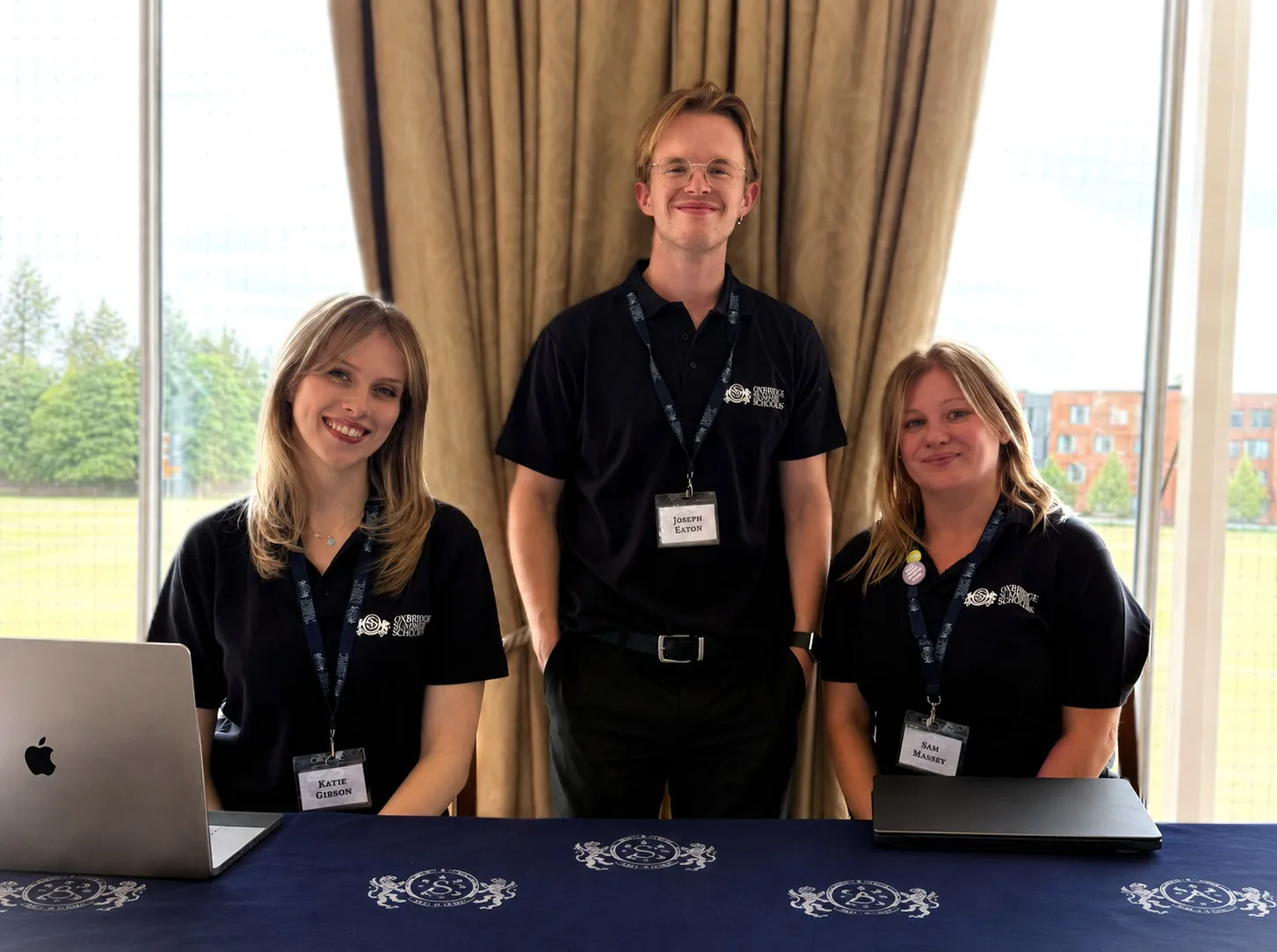 Three people wearing black Oxford Summer Schools polo shirts with name tags sit and stand behind a table with laptops, in front of a window with curtains and outdoor view.