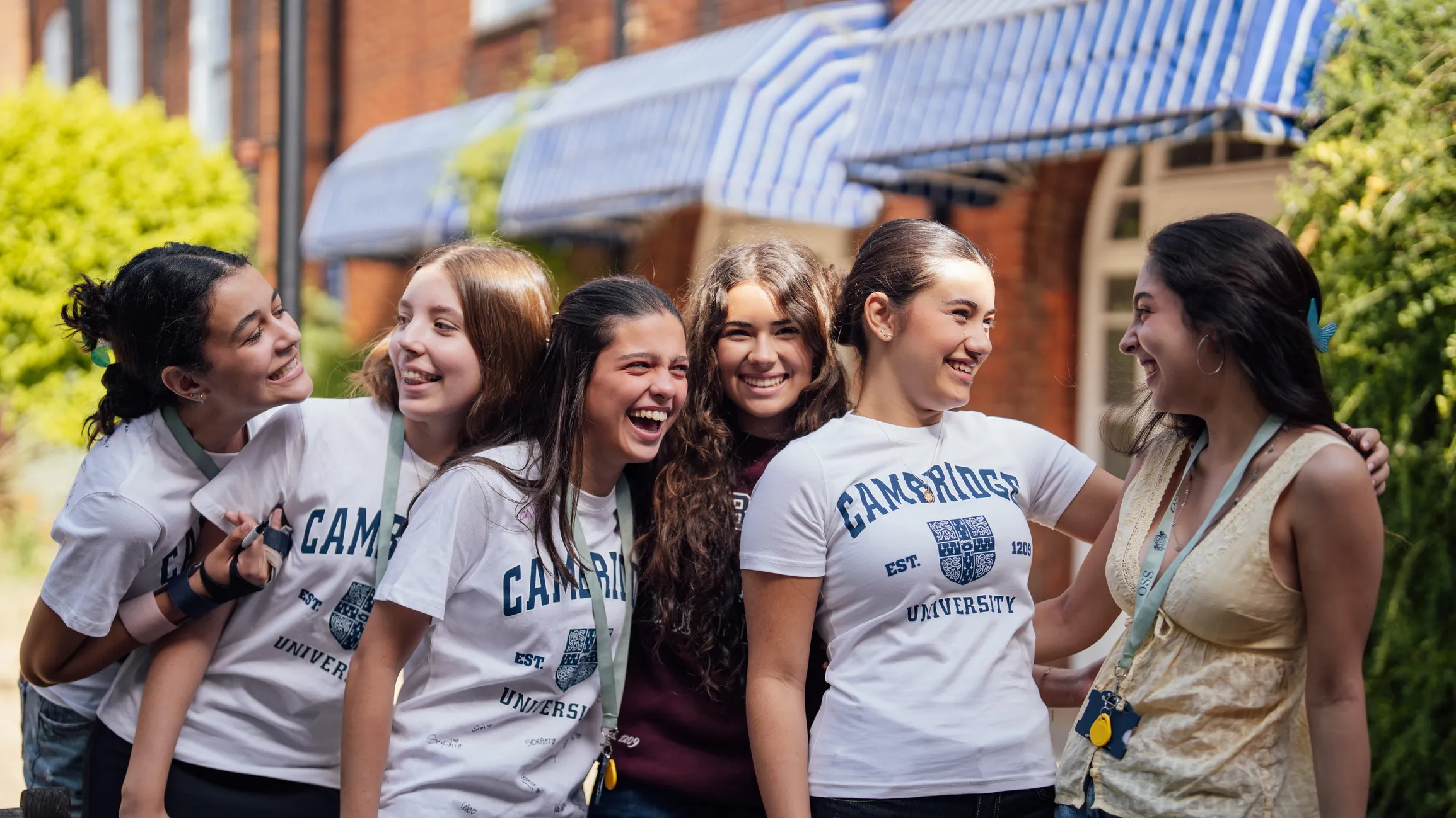 Group of six young women smiling and laughing outdoors with greenery and blue-striped awnings on a brick building behind them.