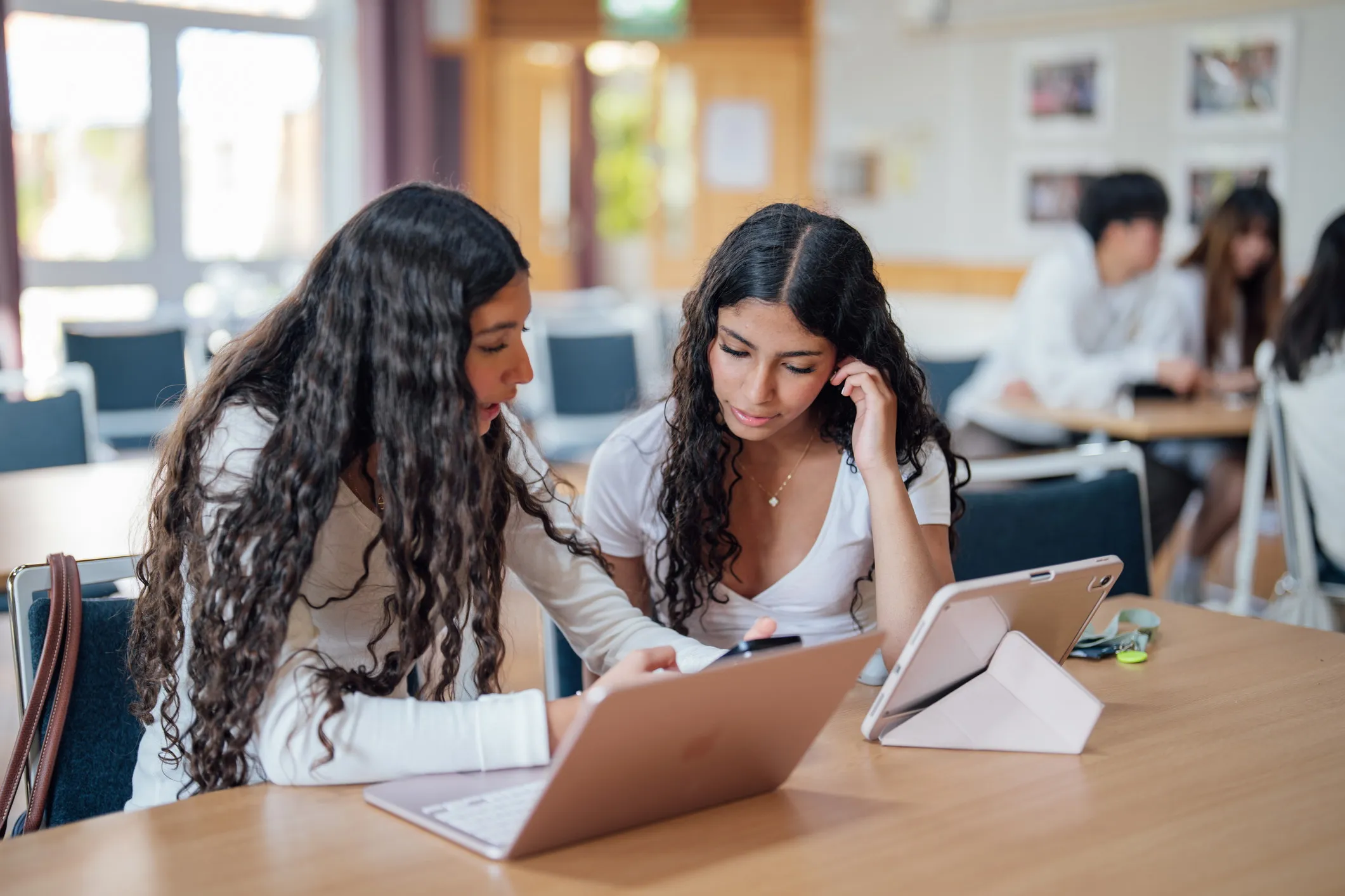 Two young women with long curly hair sitting at a wooden table, focused on a smartphone and using a laptop and a tablet in a classroom setting.