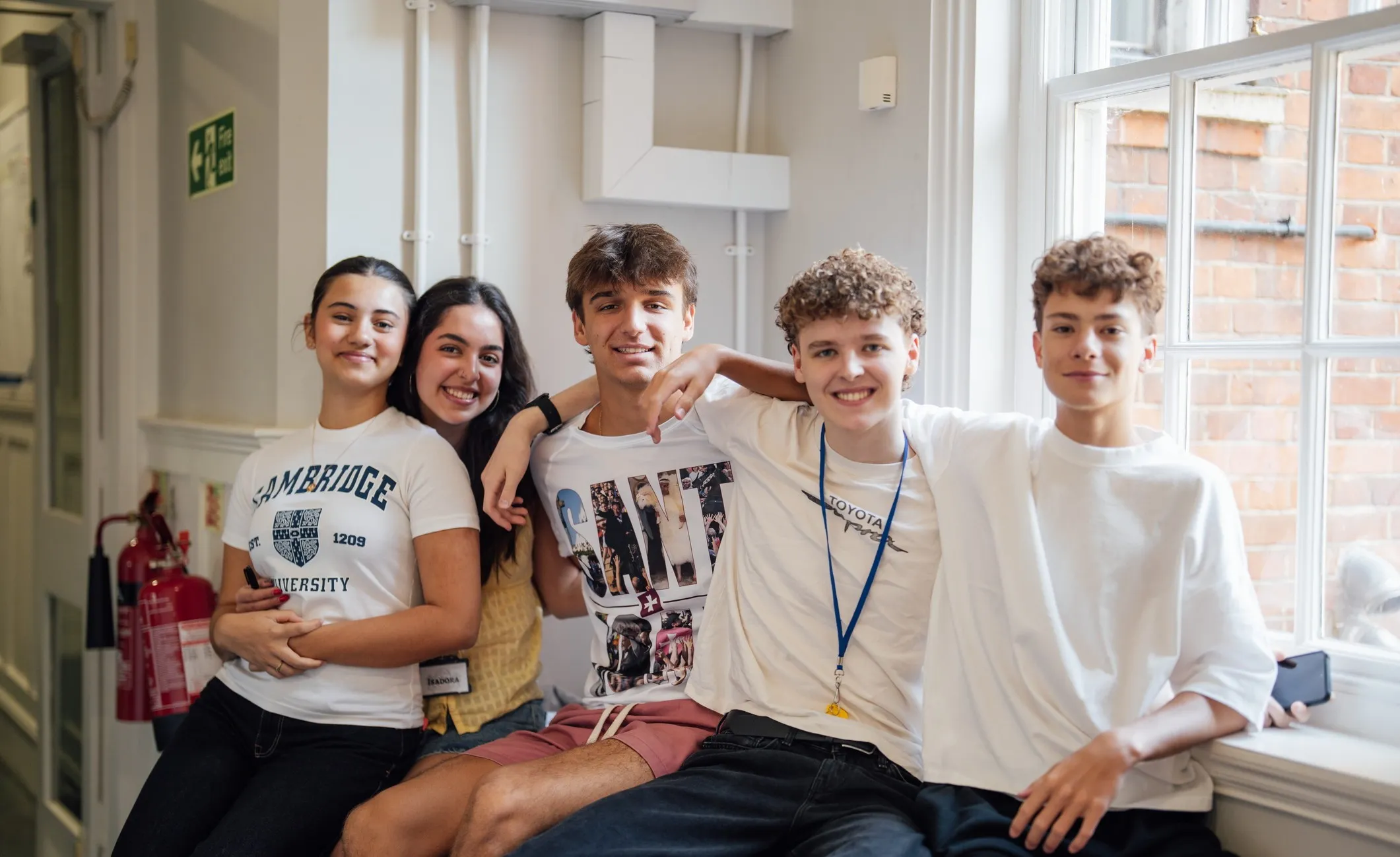 Five smiling young people sitting closely together indoors near a window, posing for a group photo with casual clothing.