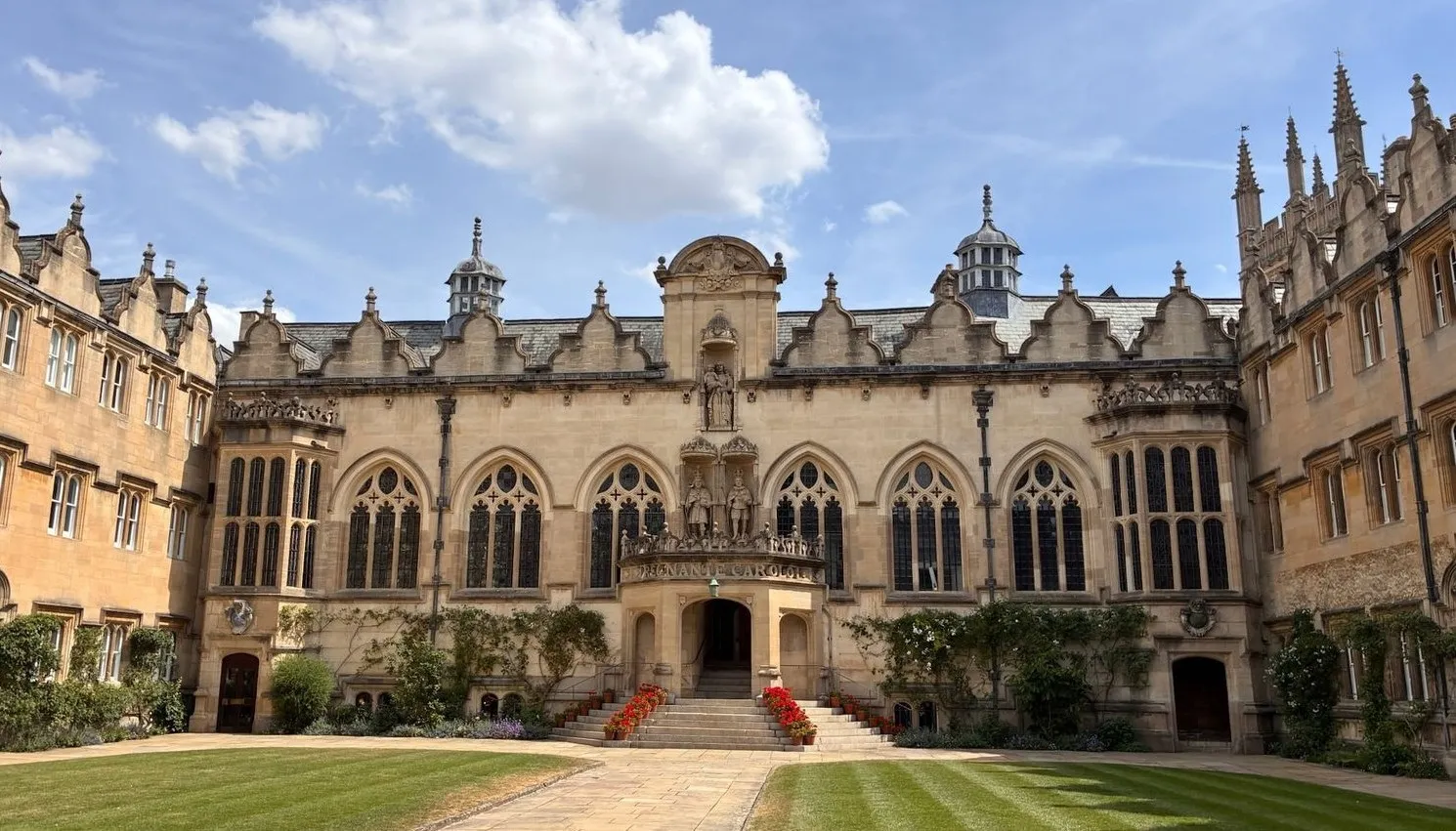 Historic stone building with ornate windows, statues above central arched entrance, and symmetrical towers under a partly cloudy sky, surrounded by manicured green lawns.