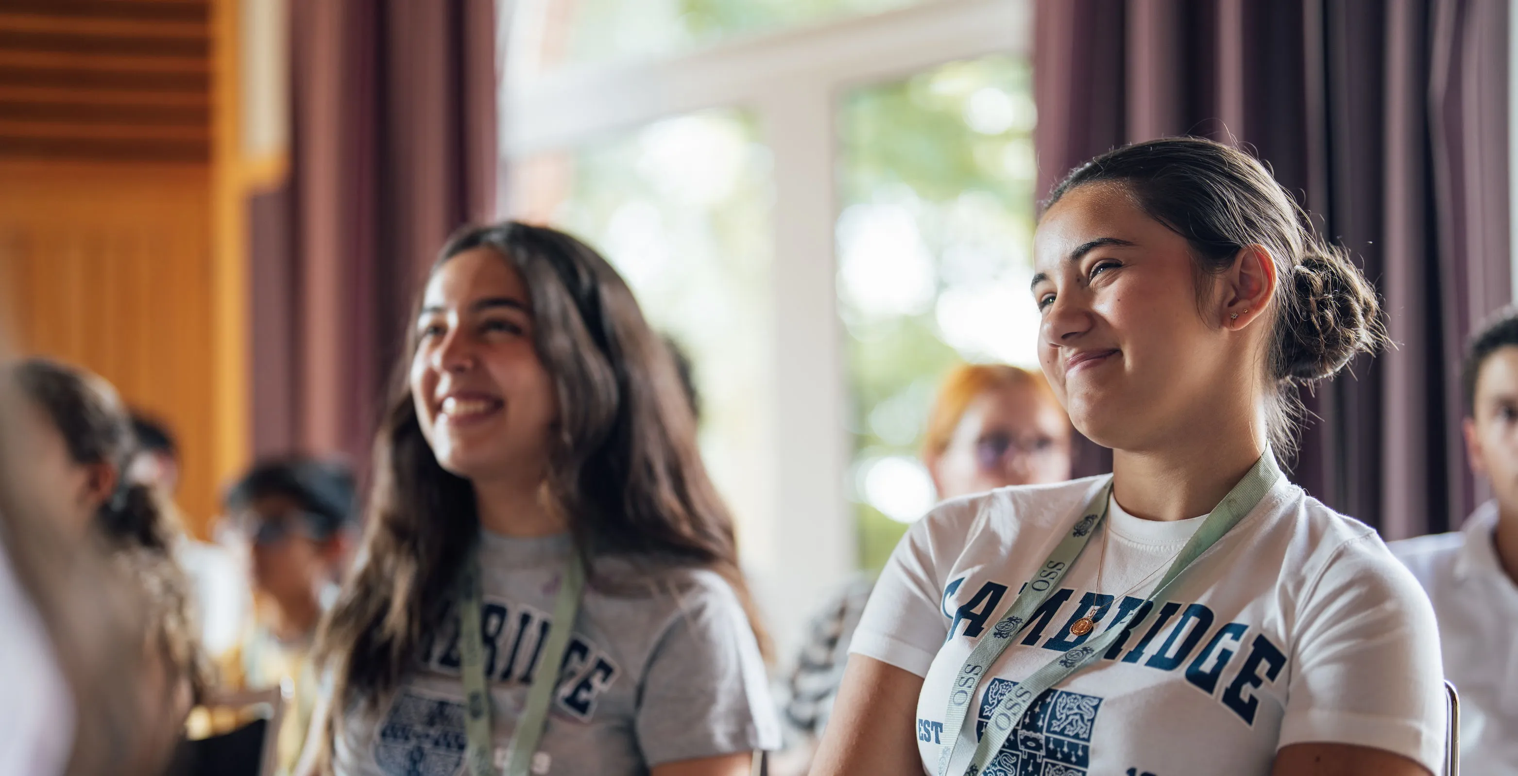 Two young women smiling and sitting in a classroom or seminar setting, wearing lanyards and casual clothing with blurred attendees and windows in the background.