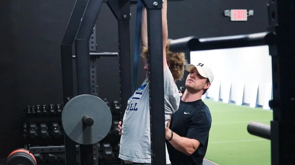 Trainer assisting a young man performing pull-ups on a bar in a gym with weightlifting equipment.