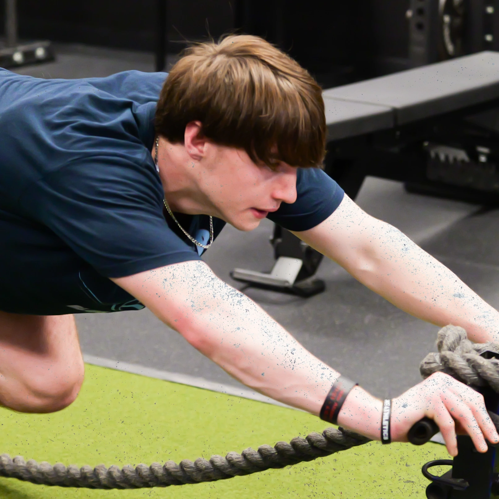 Young man in a gym wearing a dark shirt pulling a weighted sled with thick ropes.