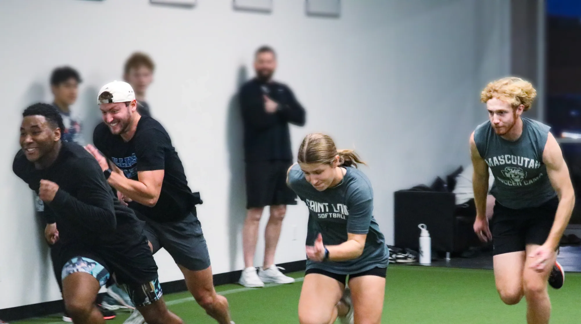 Group of young adults sprinting indoors on green turf during a fitness training session.