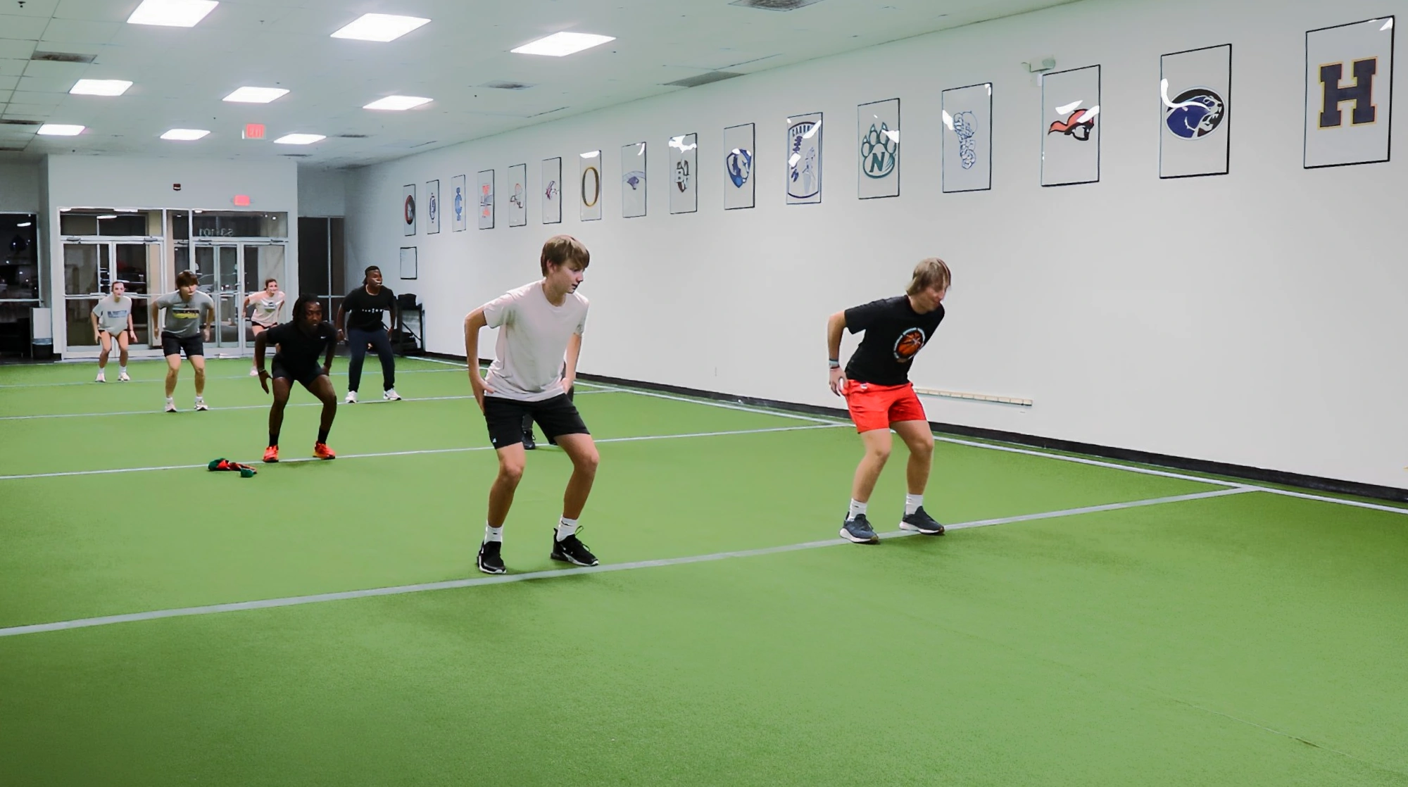 Six young athletes indoors on artificial turf, performing a squat or jumping exercise near a white wall with framed sports logos.