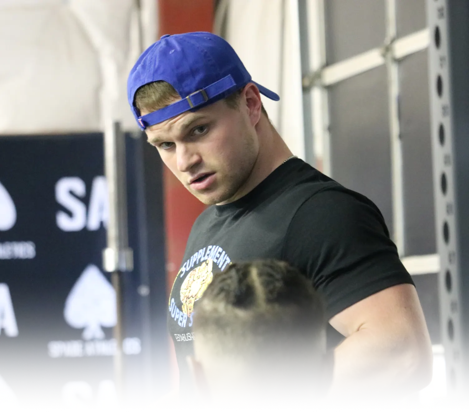 Man wearing a blue backward cap and black t-shirt looking intently at another person in a gym setting.