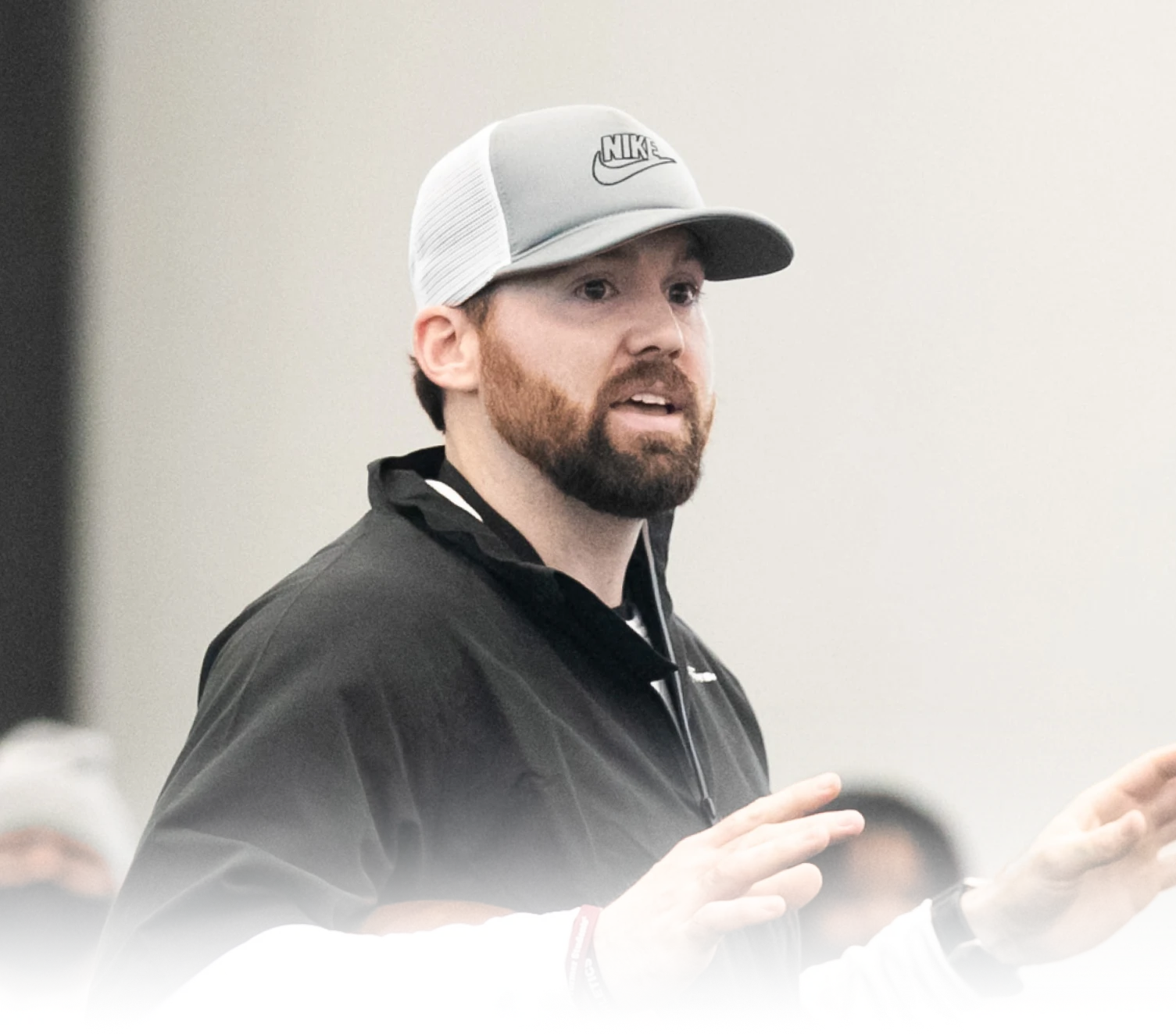 Man with a beard wearing a gray Nike cap and black jacket speaking with hands raised in a gesture.