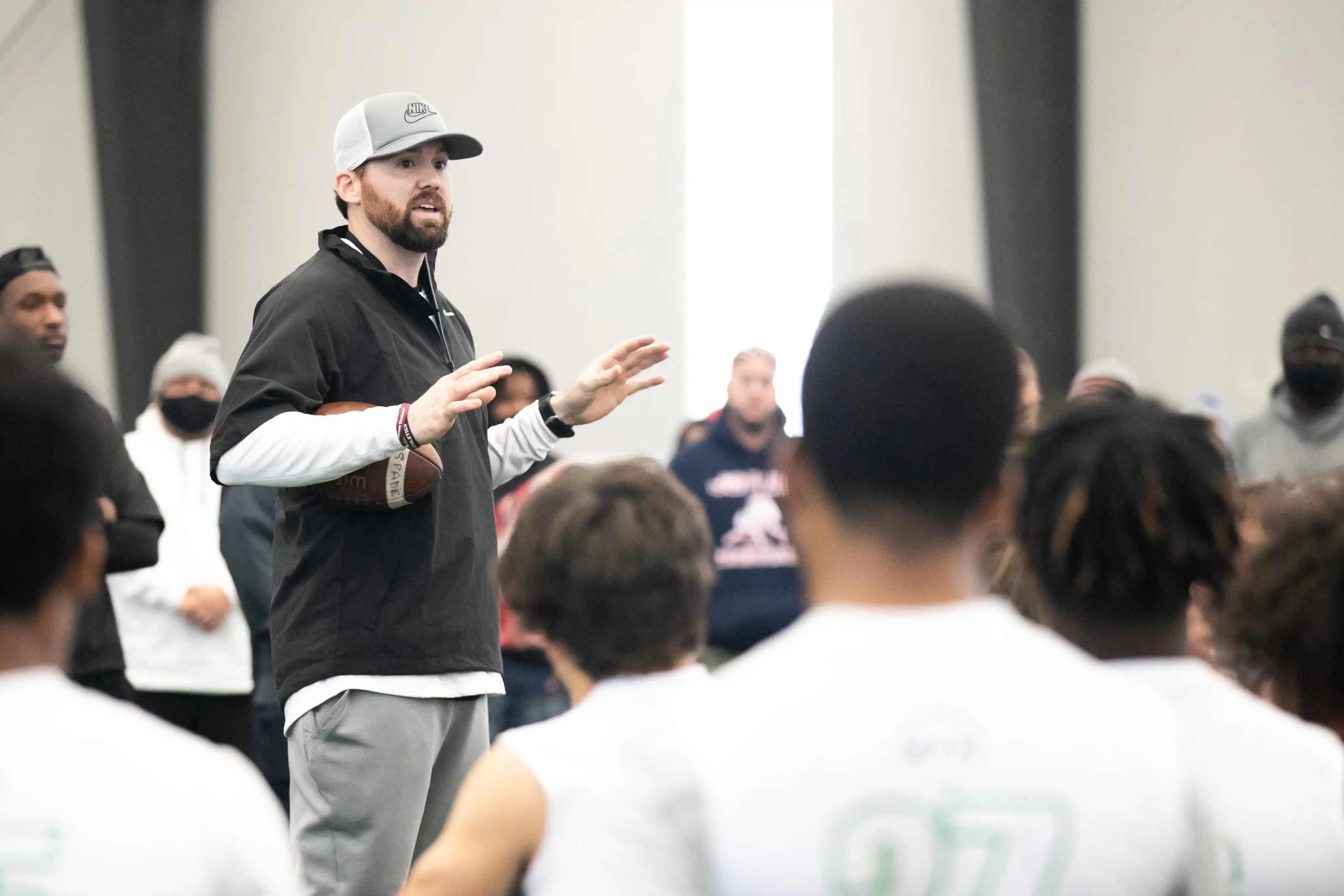 Coach wearing a gray Nike cap holding a football and speaking to a group of athletes in white shirts.