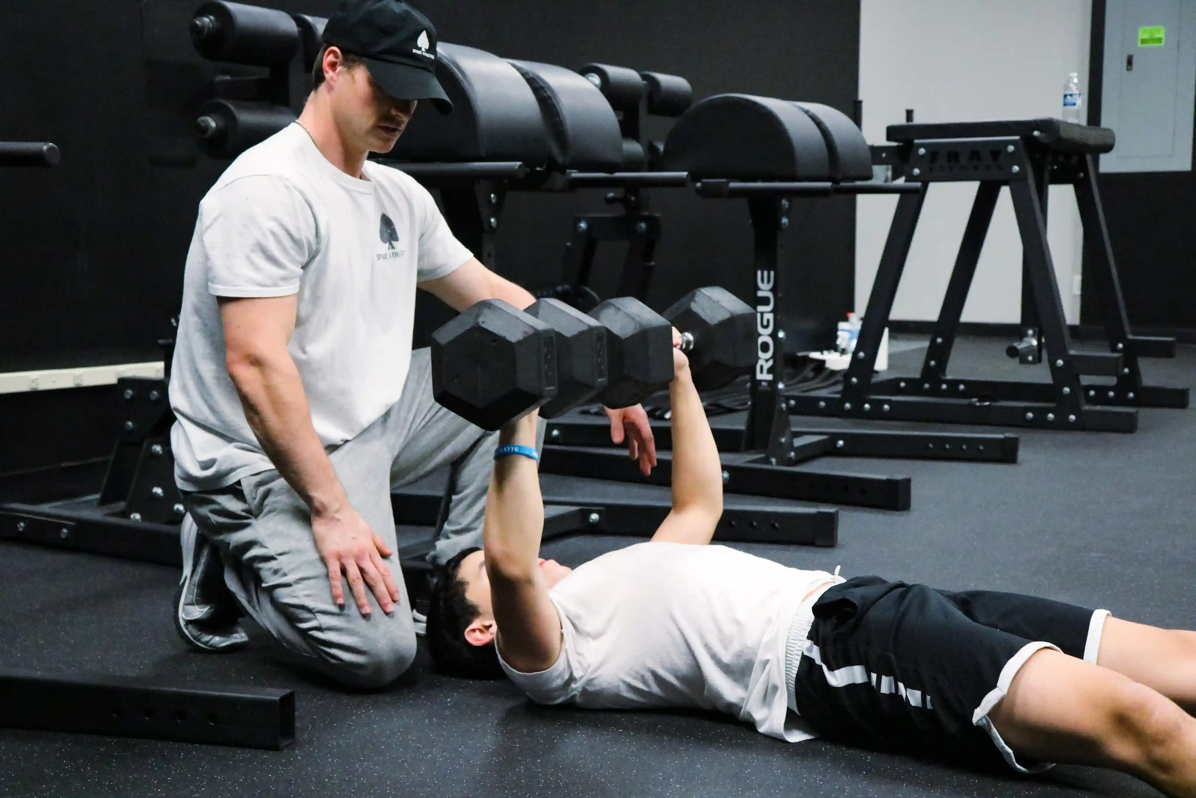A fitness trainer kneeling next to a man lying on the gym floor lifting dumbbells during a workout.