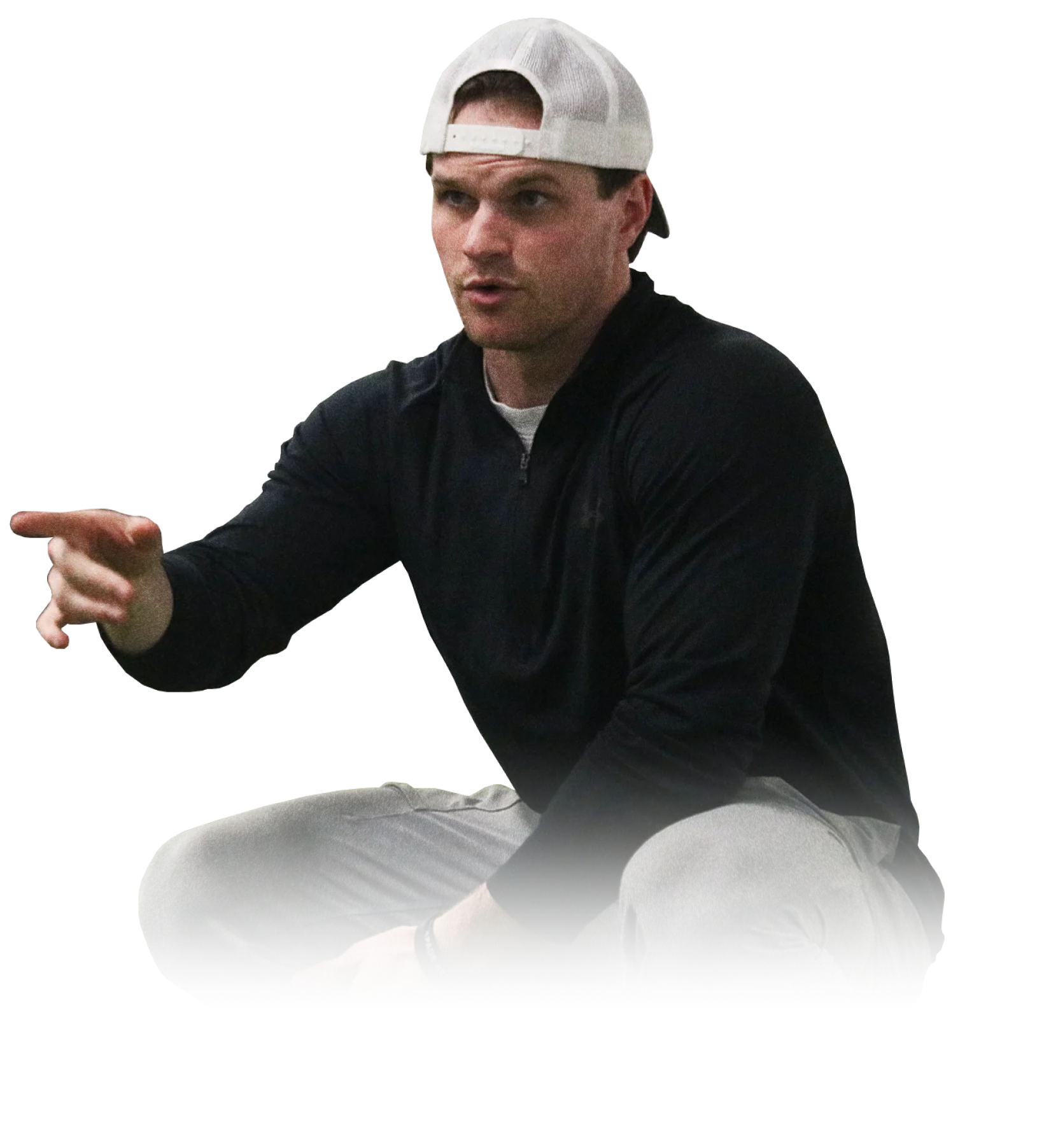 Man wearing a backwards white cap, black long-sleeve shirt, and gray pants squatting and pointing with his right hand.