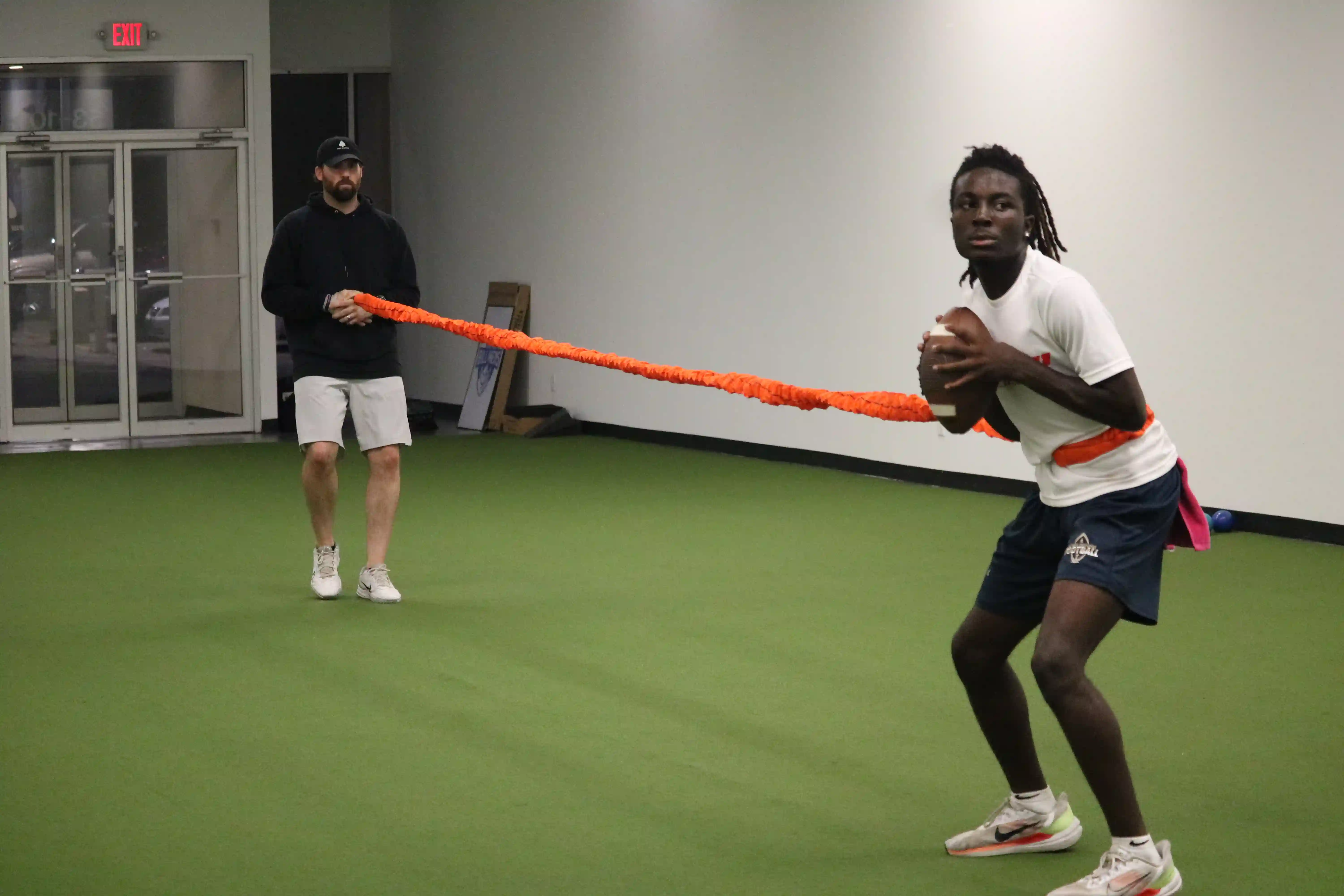 Two men indoors, one wearing resistance training gear with a football preparing to throw, the other holding the resistance band behind him.