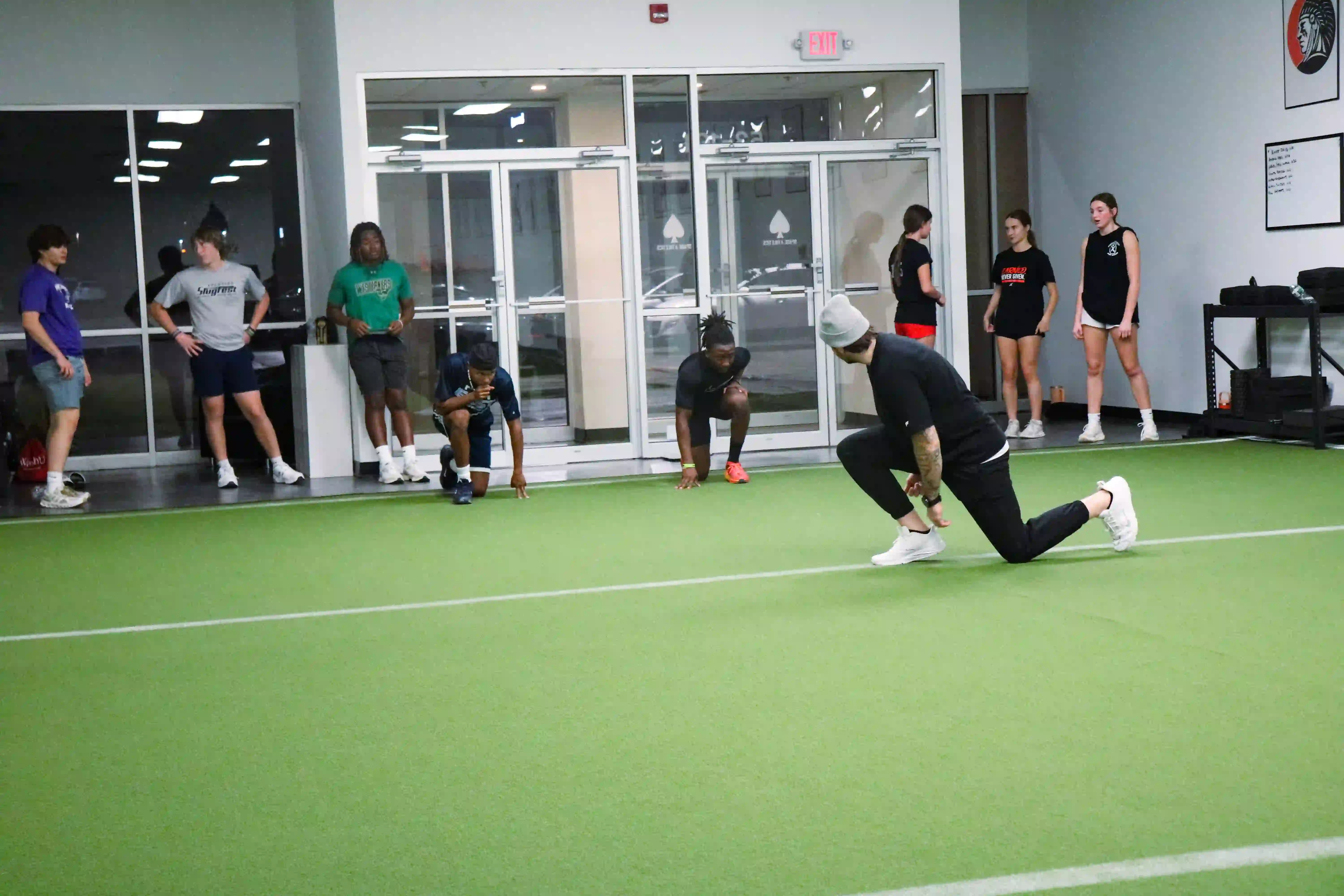 Group of young people in athletic clothing indoors on artificial turf, some preparing to sprint while others stand and watch.