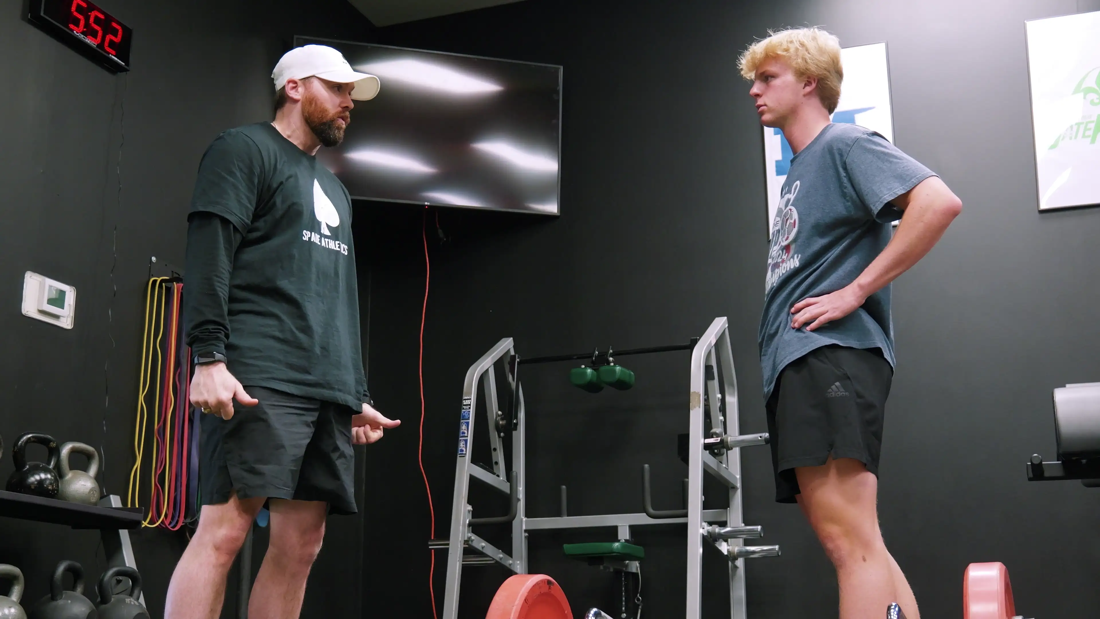Two men in a gym facing each other near weightlifting equipment, one wearing a white cap and black shirt, the other in a gray shirt.