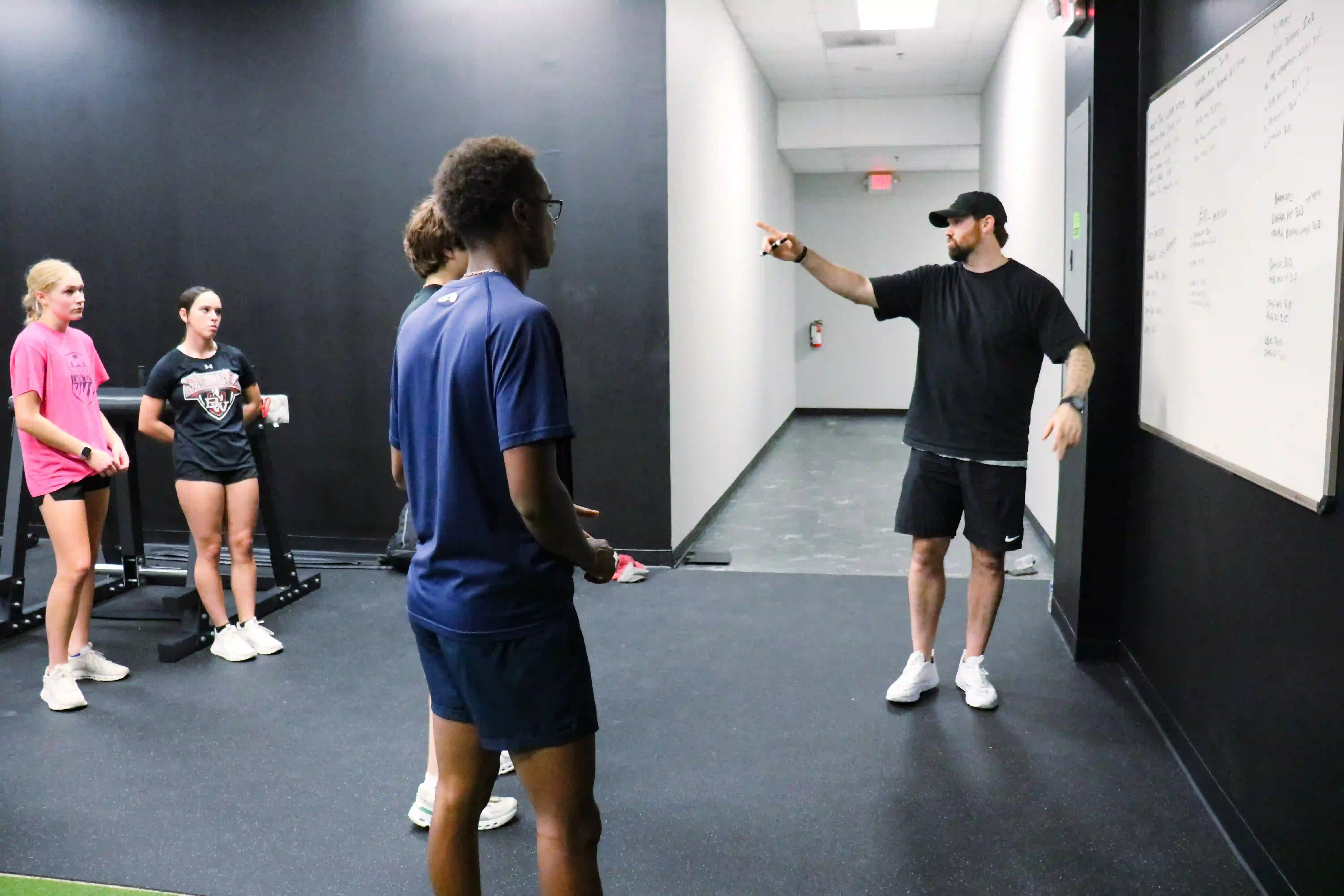 Coach in black outfit pointing and instructing a group of young athletes in a gym hallway.