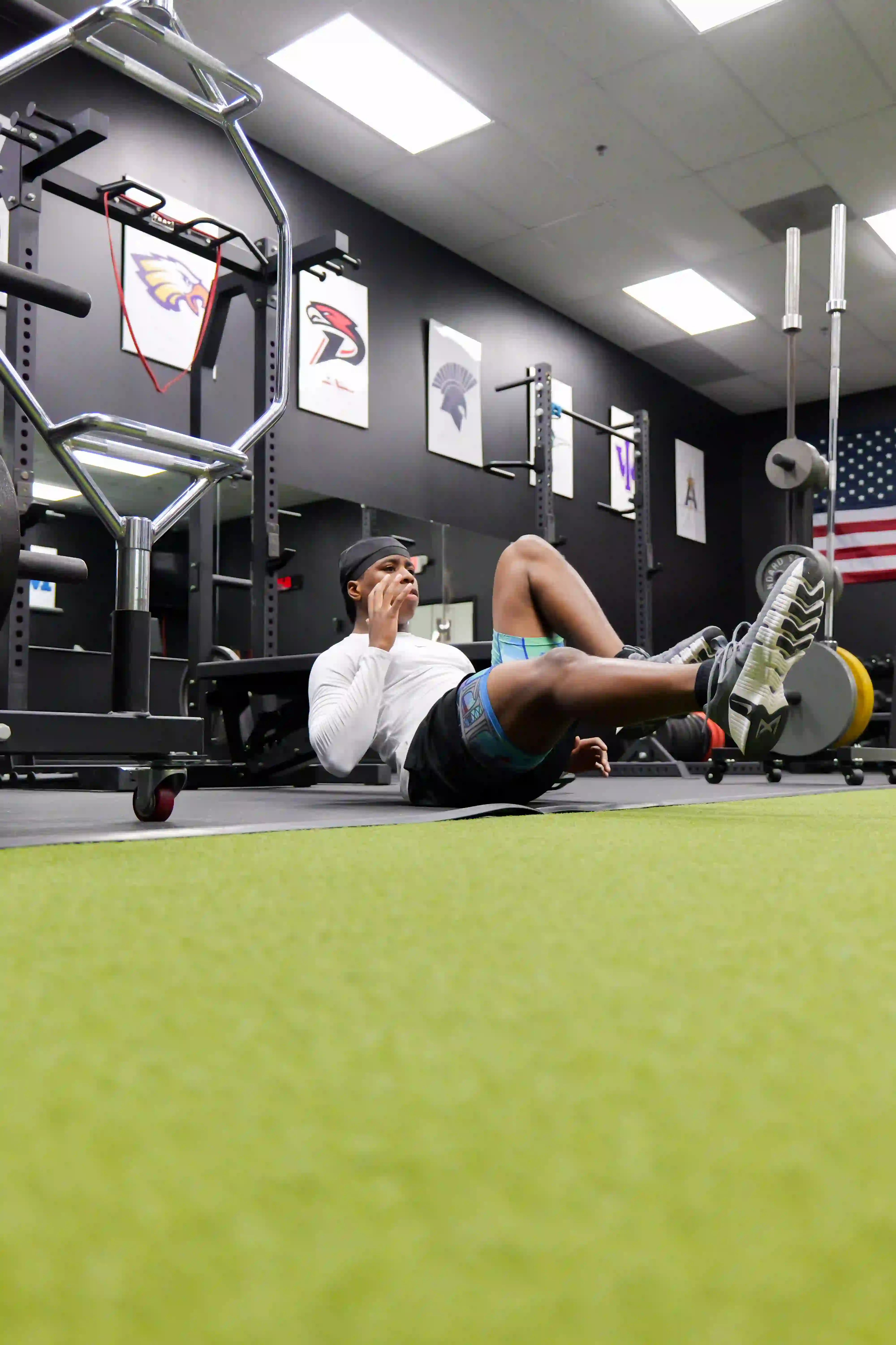 Young man exercising on the floor in a gym with weightlifting equipment and an American flag in the background.