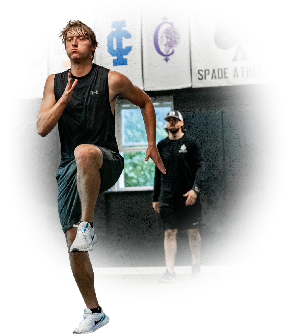 Athlete in black sleeveless shirt and gray shorts sprinting indoors with a coach observing in the background.