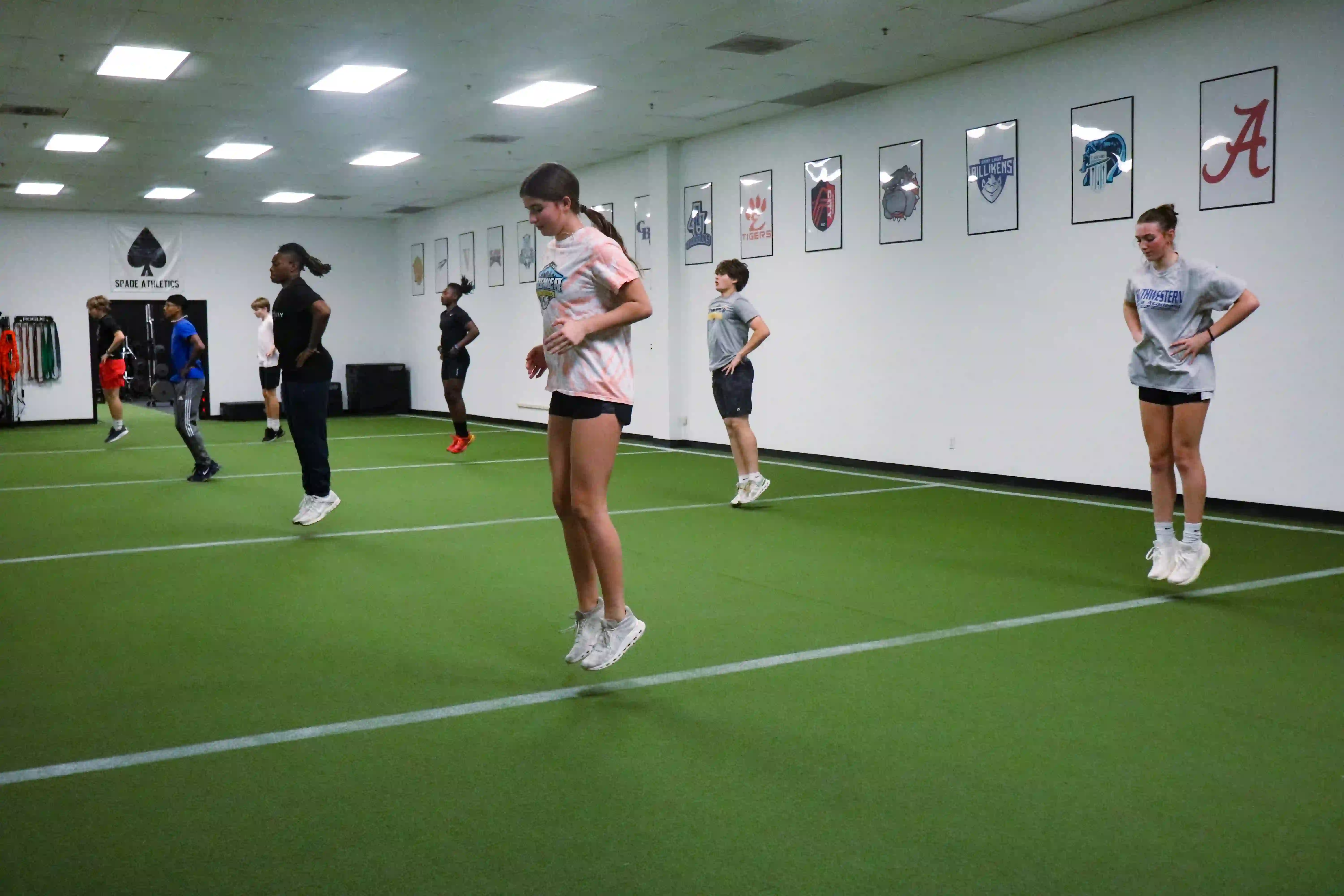 Group of young athletes performing jump exercises inside a gym with green turf and sports banners on the wall.