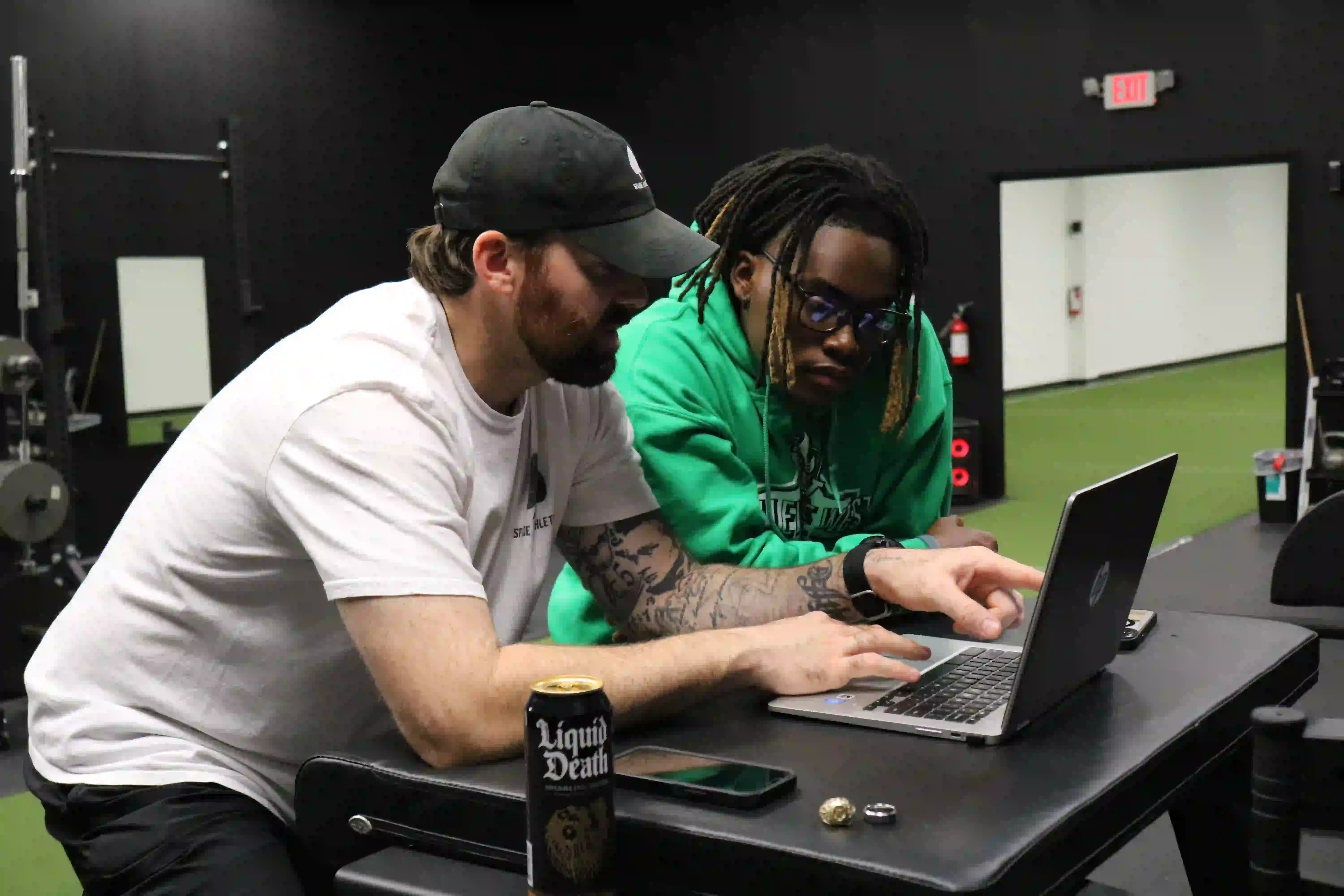 Two men sitting at a table in a gym, one wearing a white t-shirt and cap pointing at a laptop screen while the other in a green hoodie looks on.
