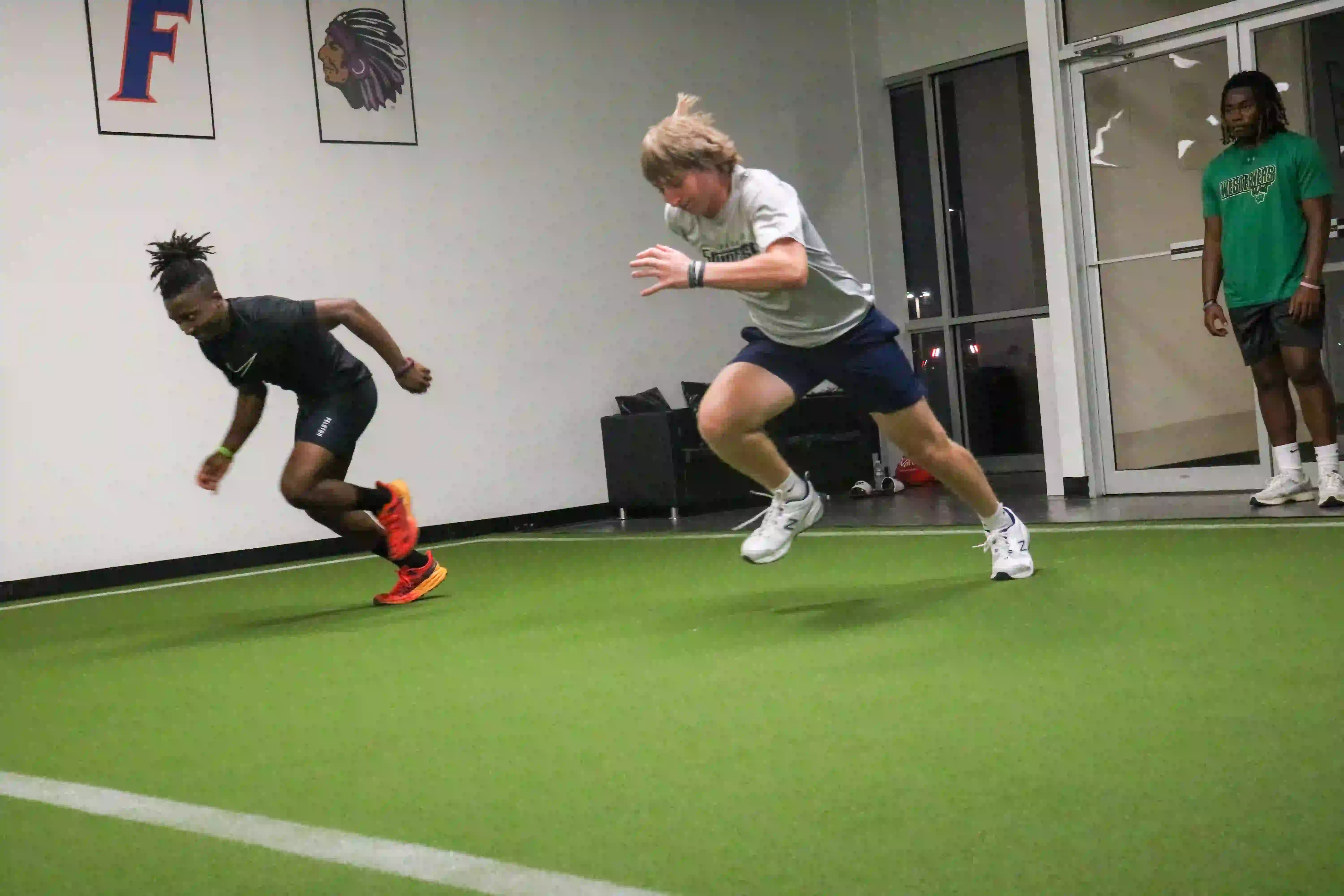 Two athletes sprinting indoors on green turf while another person watches near a glass door.