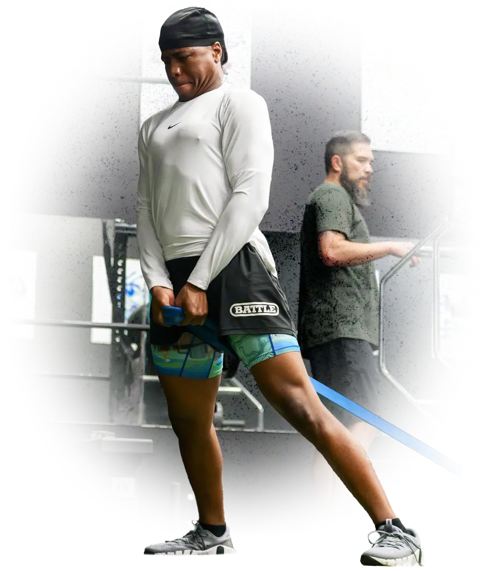 Athlete performing resistance band exercise in gym with a trainer observing in the background.