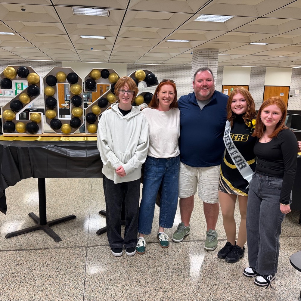 Five people smiling indoors near a table decorated with black and gold balloons shaped into the numbers 2023.