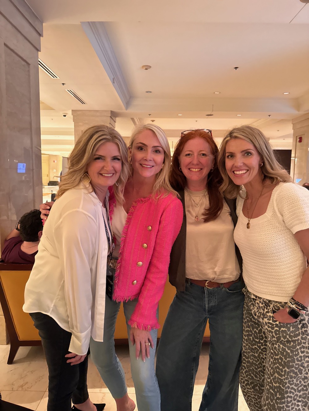 Four smiling women standing close together in a warmly lit indoor setting, wearing casual and business-casual attire.