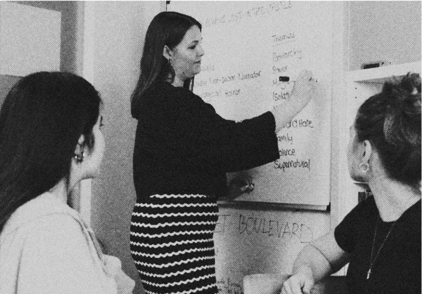 A woman writing on a whiteboard while two other women watch attentively in a meeting or classroom setting.