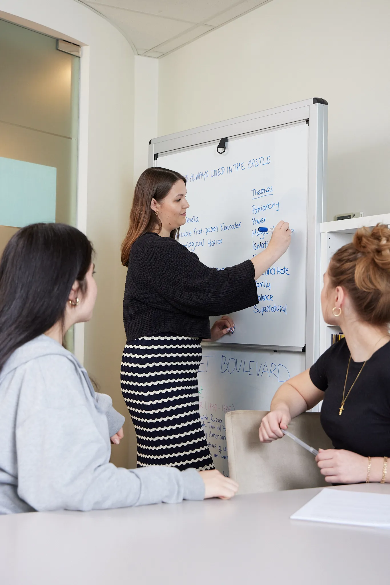 Woman writing blue text on a whiteboard while two others watch, seated at a table in a classroom.