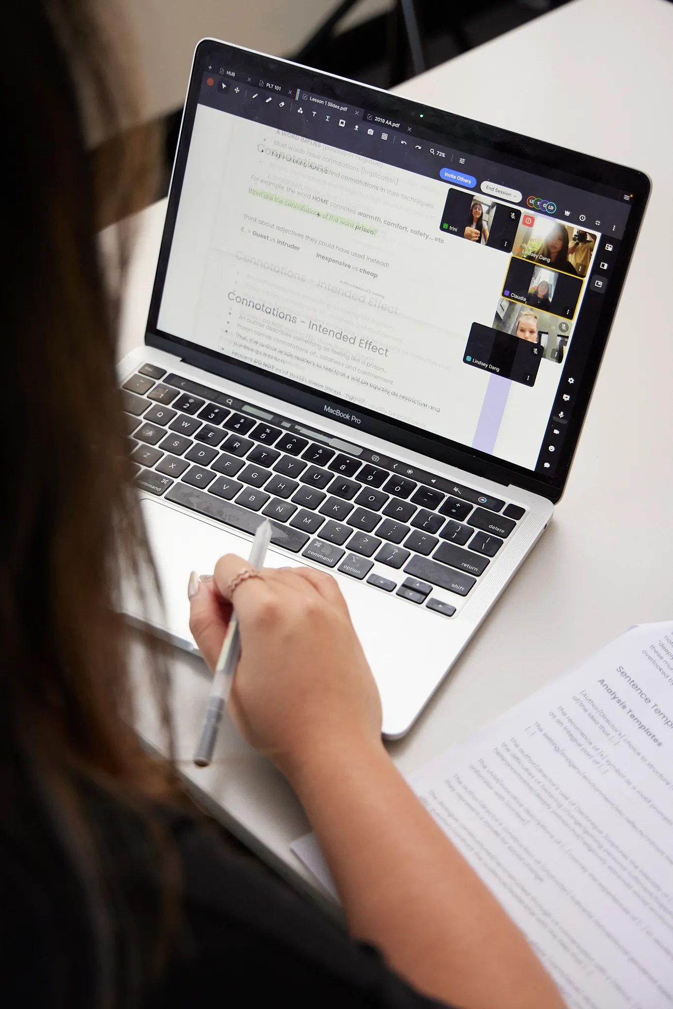 Person attending an online video conference on a MacBook Pro while taking notes with a pen over printed documents.