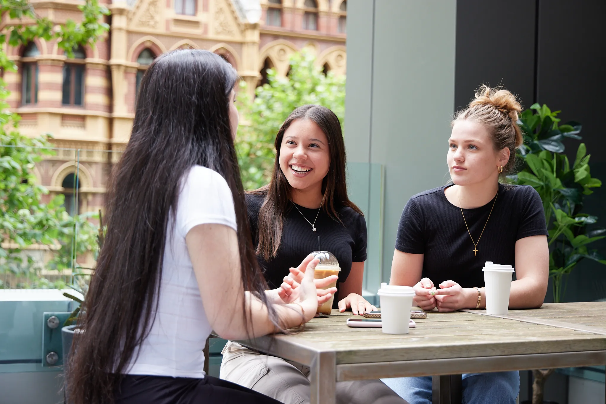 Three young women sitting at an outdoor wooden table, chatting and drinking coffee, with a historic building and greenery in the background.