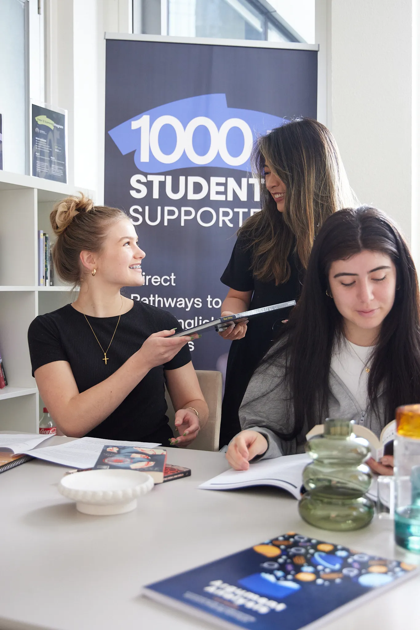 Three students in a study room; one sitting and smiling while handing a book to another standing student, with a third student reading nearby.