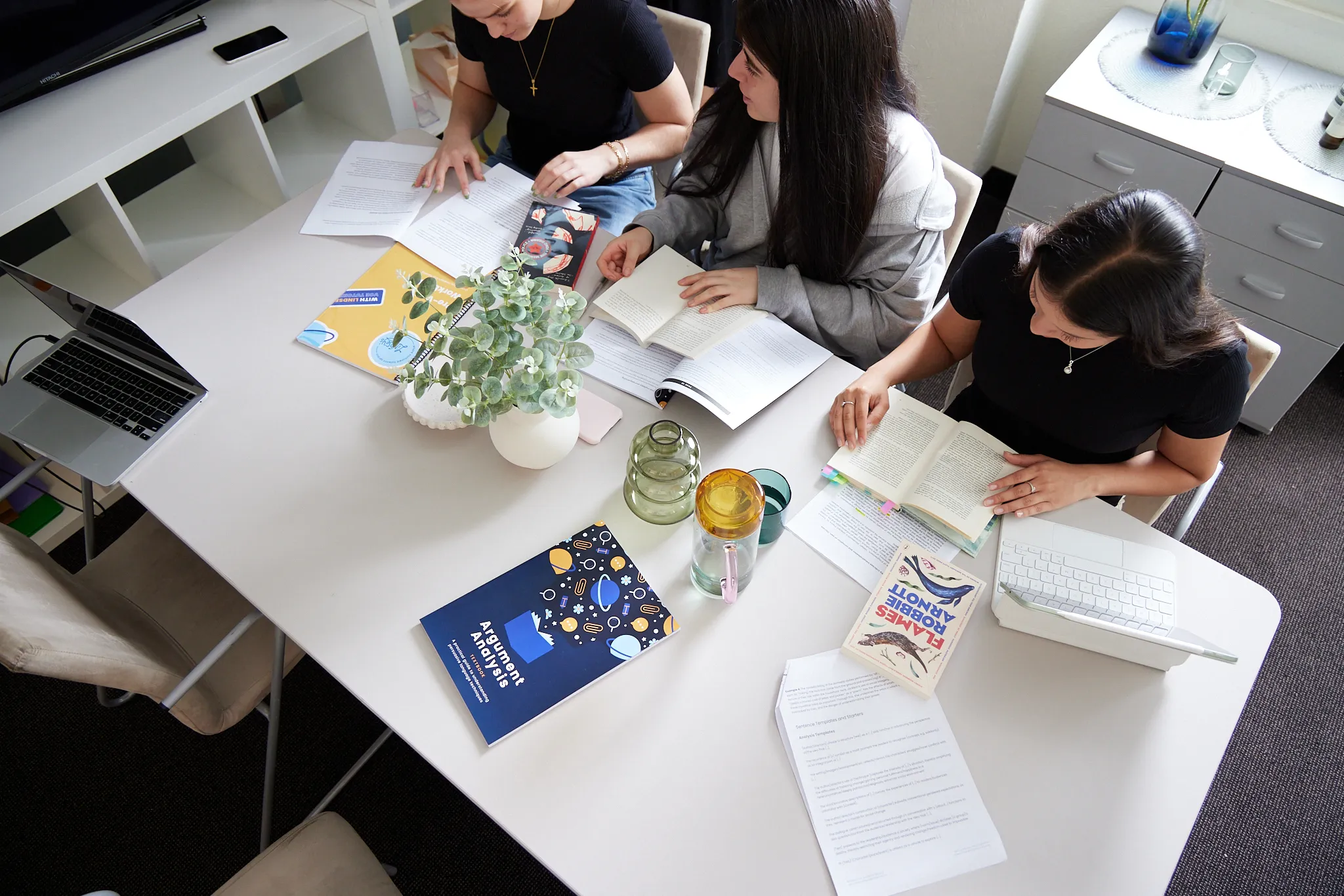 Three people sitting around a table reading books and papers with a laptop, plant, and water glasses on the table.