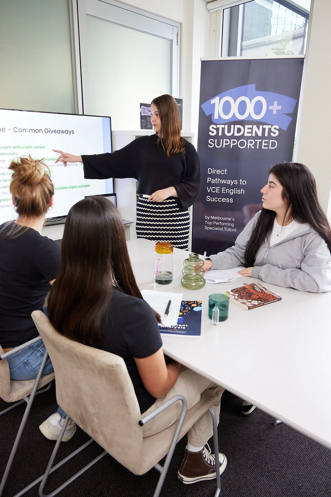 A female instructor points to a screen while teaching three female students seated around a table in a classroom setting.