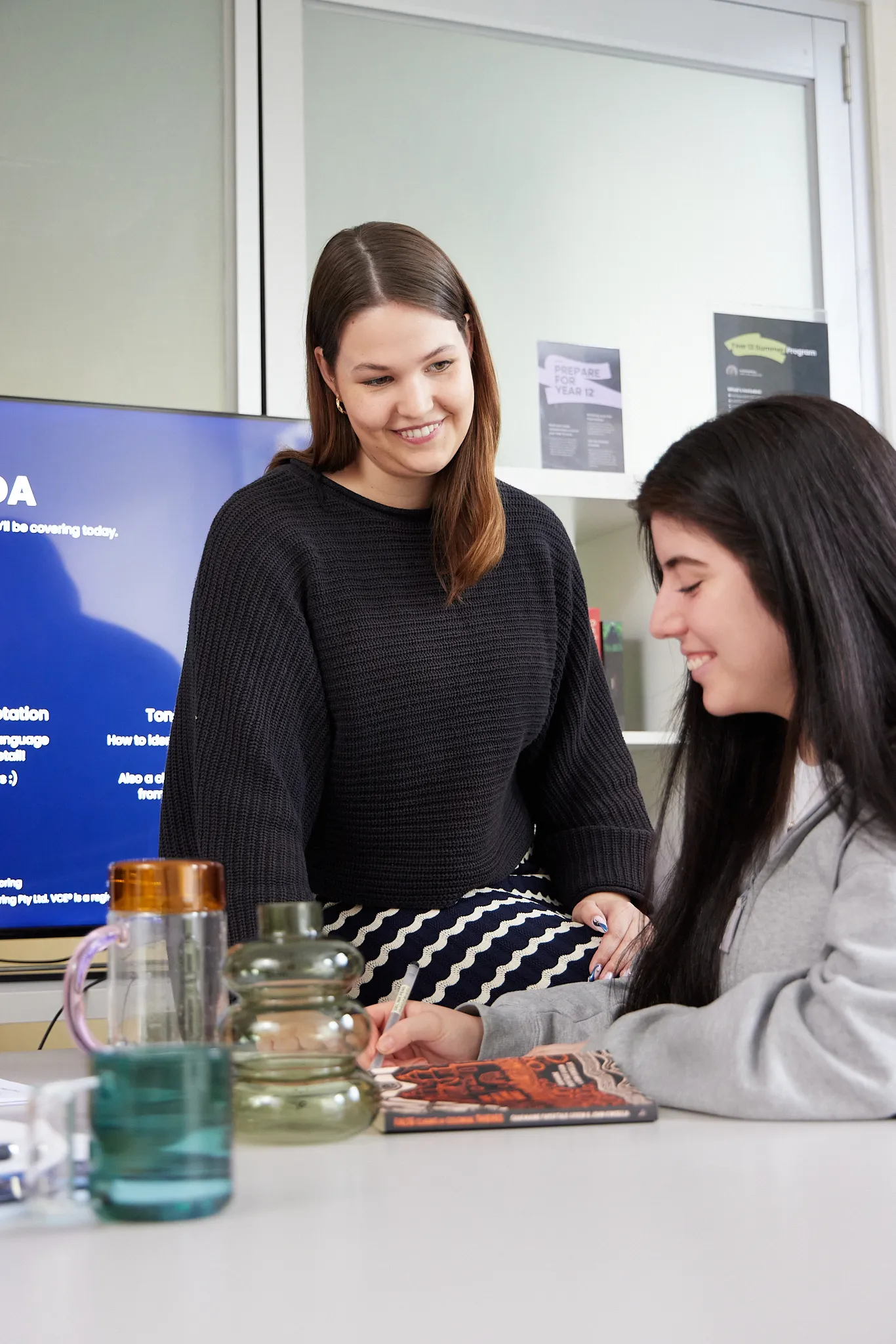 Two young women smiling and working together at a desk with books and water bottles.