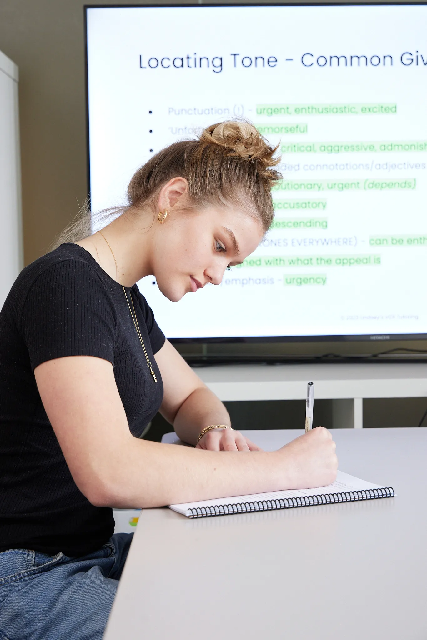 Young woman with hair bun wearing a black shirt and gold jewelry, writing in a spiral notebook at a table with a presentation slide titled 'Locating Tone - Common Giv' displayed on a screen behind her.