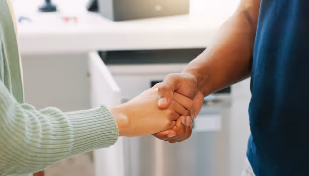 Two people shaking hands indoors, one wearing a green sweater and the other a dark blue shirt.